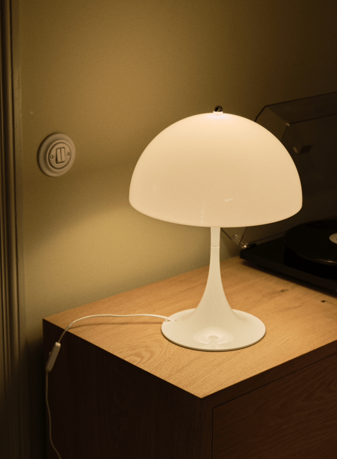 White table lamp on wooden surface beside vinyl player, © 2025 Louis Poulsen.