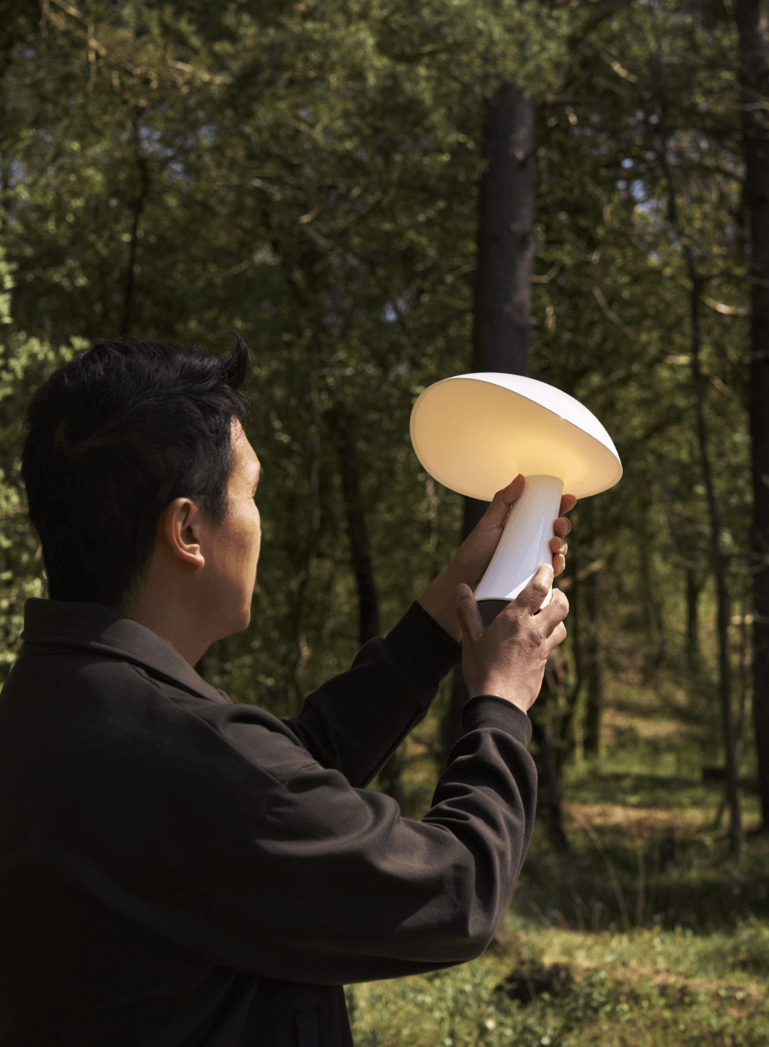 Person holding a Rumee portable lamp in a forest, with soft lighting reflecting on the surface, © 2025 Louis Poulsen.